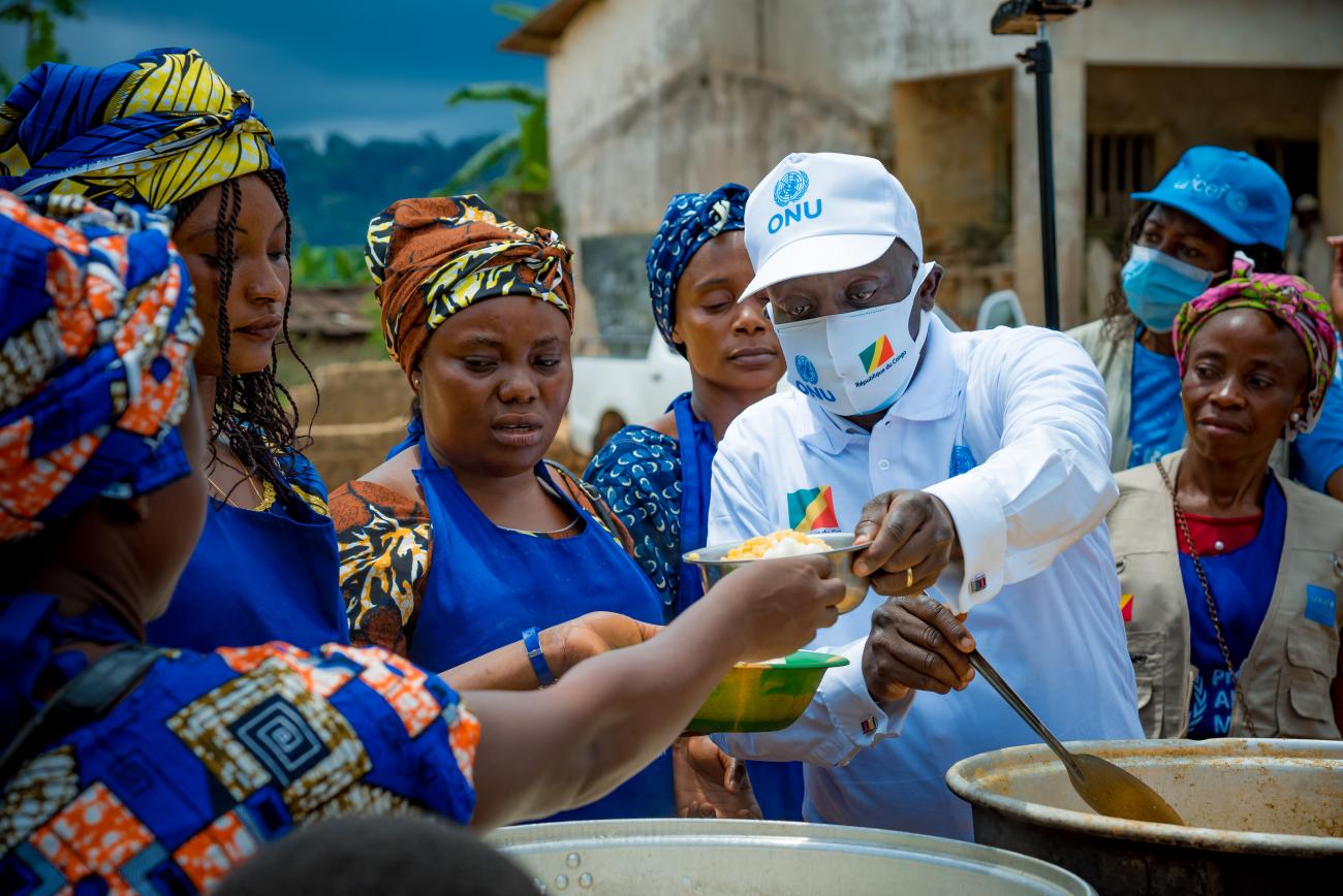 A man wearing a cap hands out food to women in blue and colored dresses. 