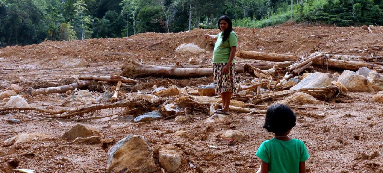 A woman in a mid-length skirt and t-shirt stands in a field of mud with tree trunk strewn around, talking to a young child.