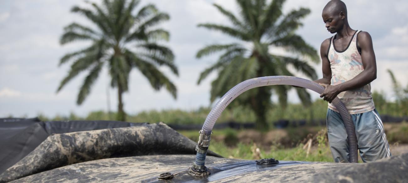 In Burundi, a young man wearing gray pants and a t-shirt without sleeves uses a hose to pump water from a portable dam set up near two palm trees.