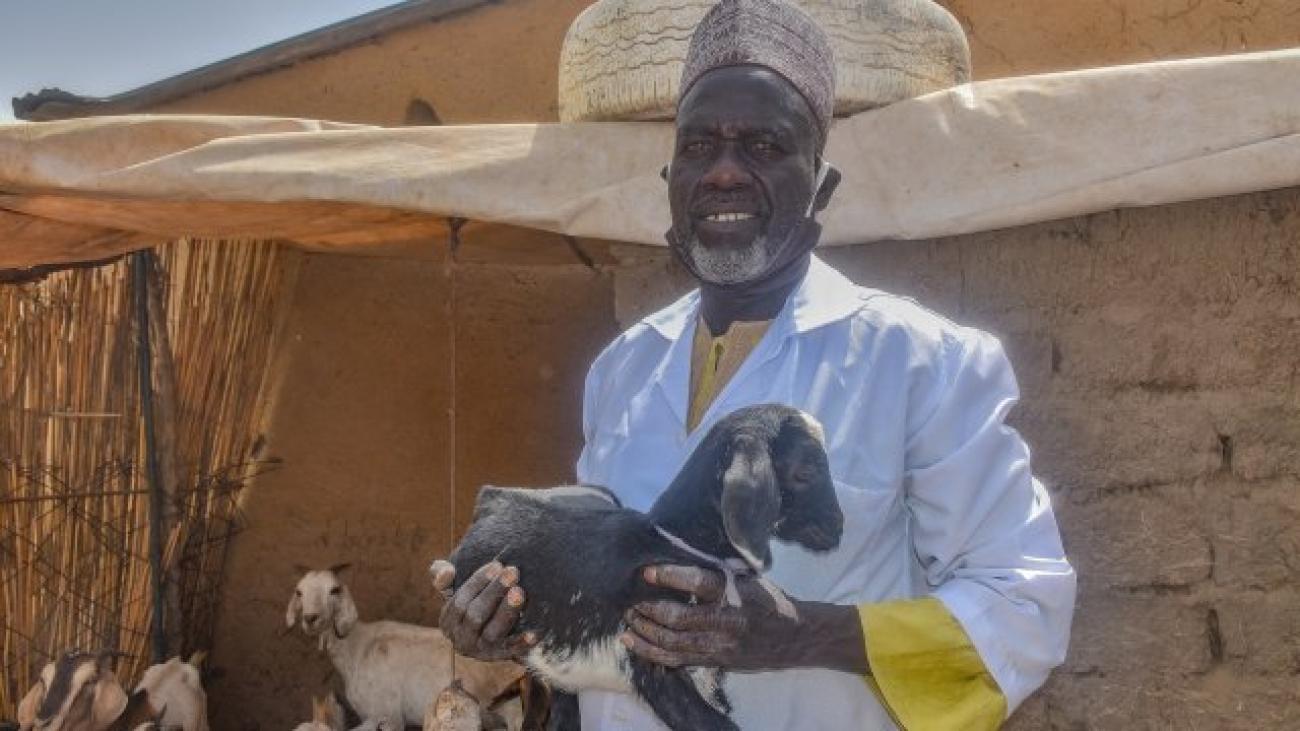 Au Cameroun, un homme portant une blouse couleur bleu ciel et un couvre-chef est photographié debout devant une maison en terre cuite, un chevreau dans les bras, face caméra.