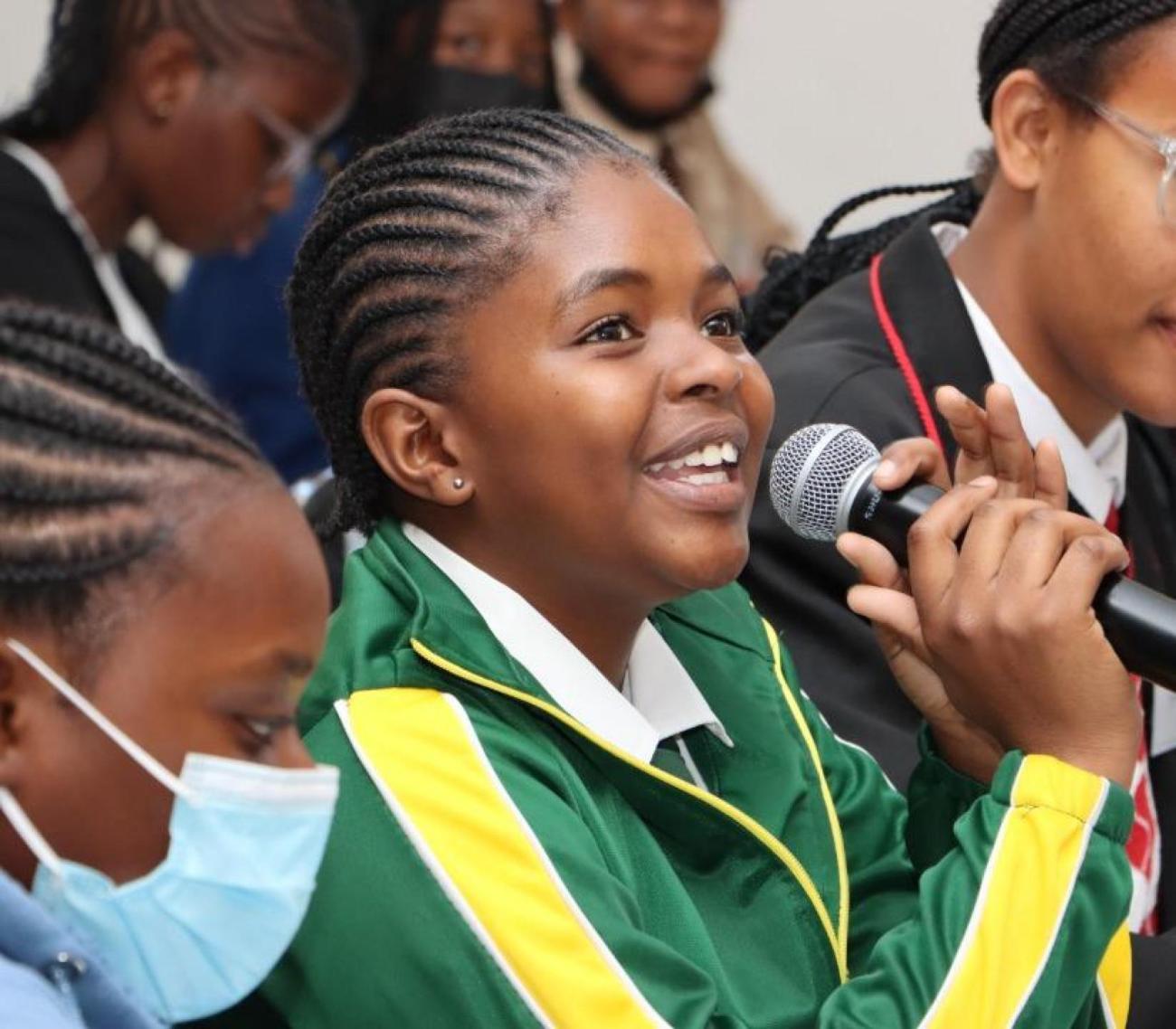 فتاة ناميبية ترتدي بدلة رياضية خضراء تجلس في غرفة مع فتيات أخريات وتتحدث في ميكروفون وهي تبتسم.