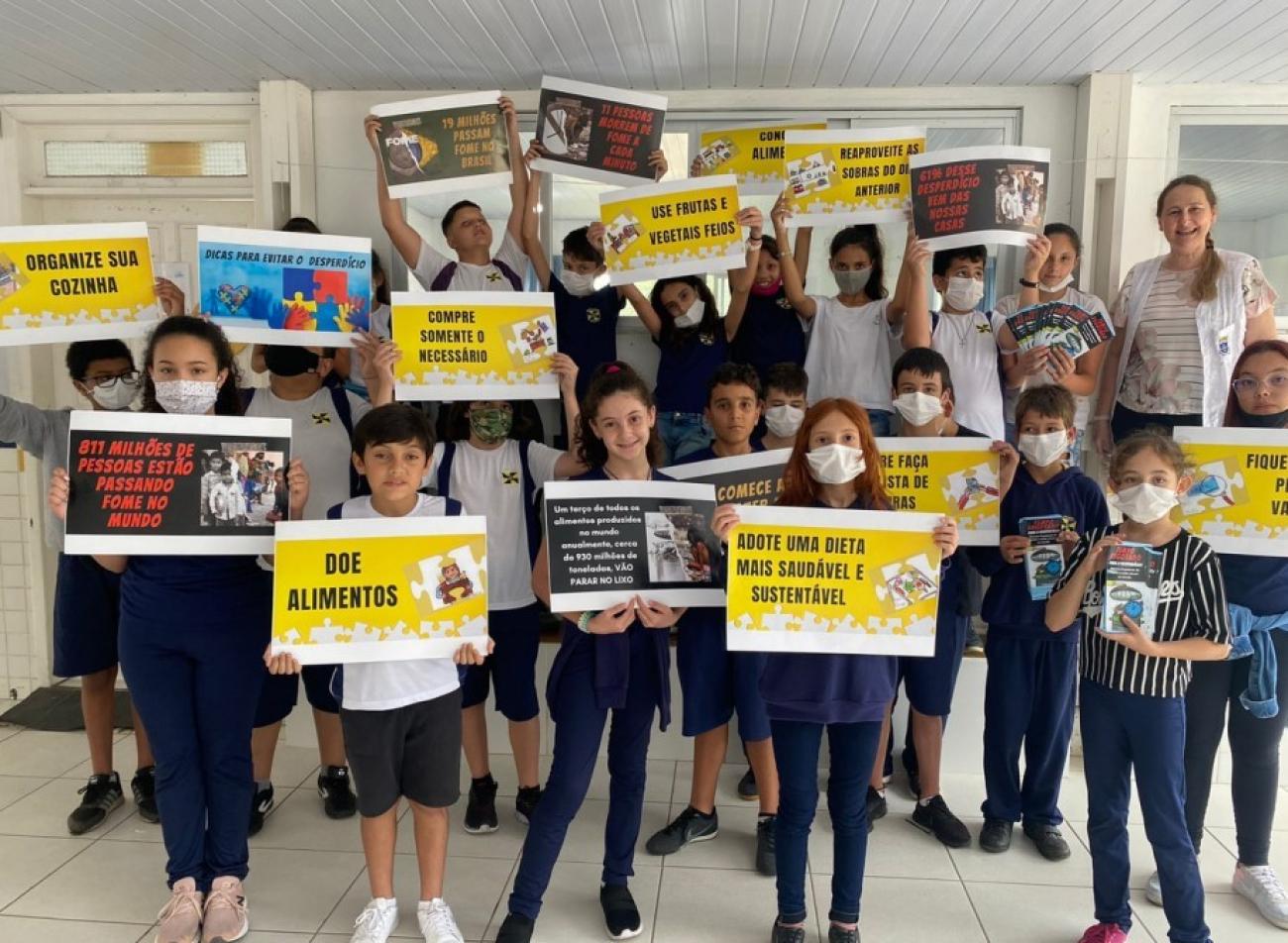 一群小学年龄的学生和他们的老师。他们都戴着口罩，站在一边，自豪地展示着他们学校关于废弃物管理的工作。