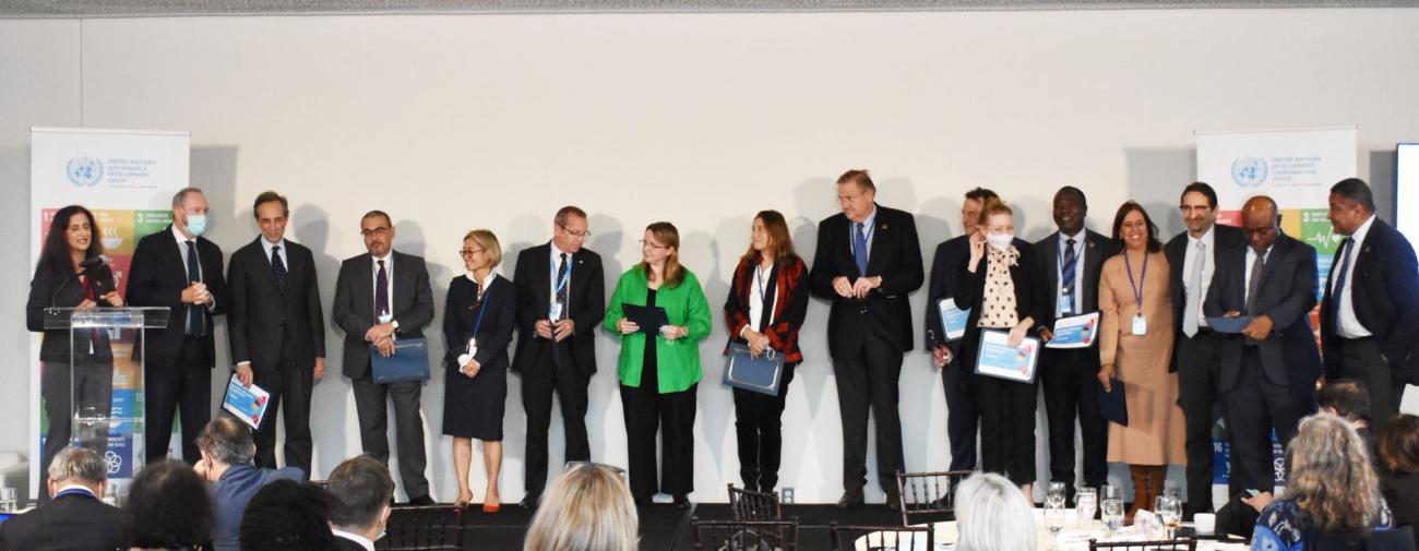 A group of sixteen people in business attire, standing next to each other, clapping and congratulating one another, on a stage during the UN Team Annual Results Reports Awards ceremony.
