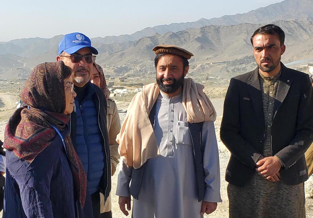 Una mujer con bufanda se encuentra junto a dos hombres con gorra y un hombre con chaqueta, con montañas de color canela en la distancia.