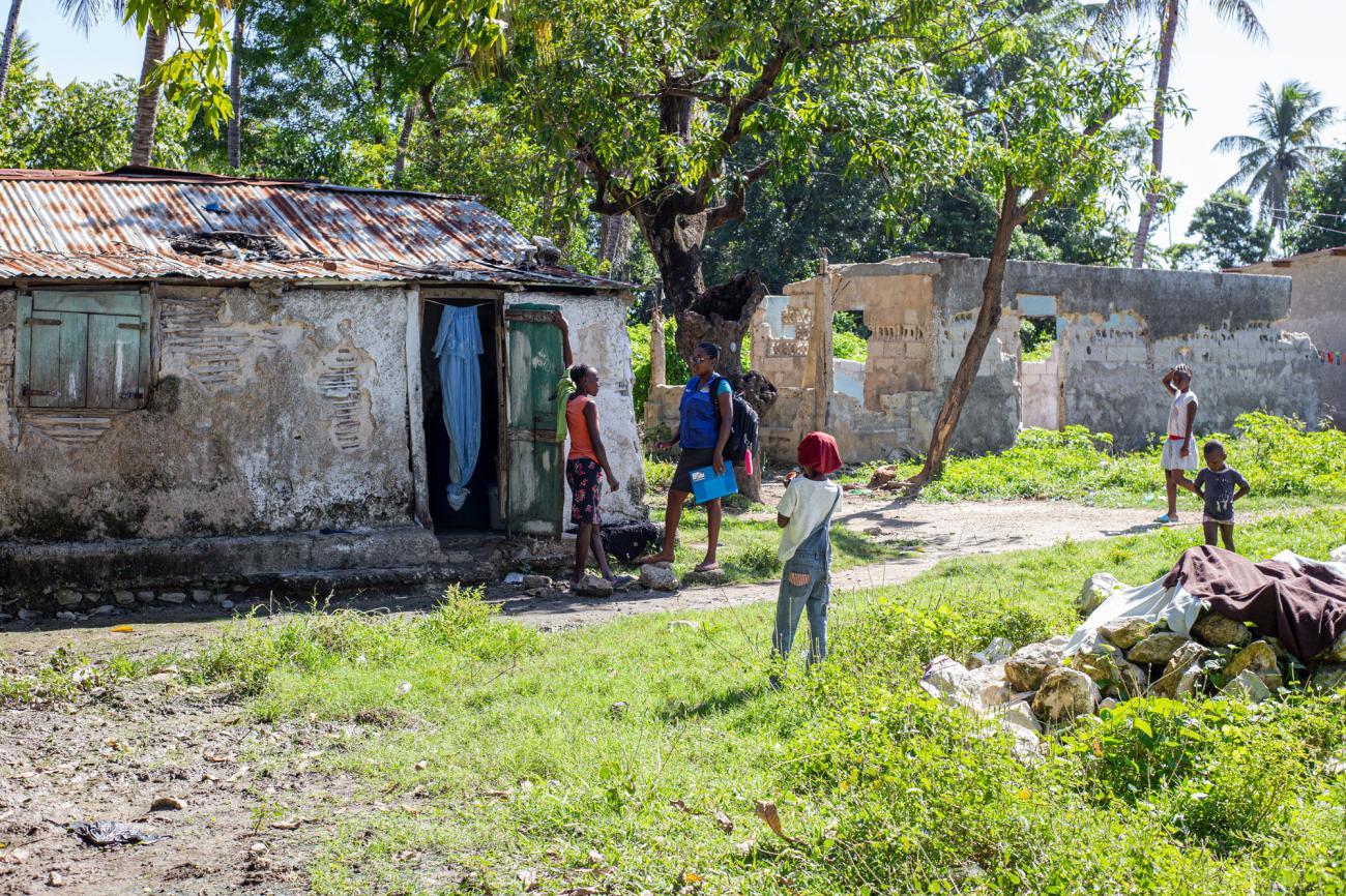 Deux jeunes femmes discutent devant l’entrée d’une vieille maison délabrée entourée d’arbres. Plusieurs enfants jouent à proximité. 