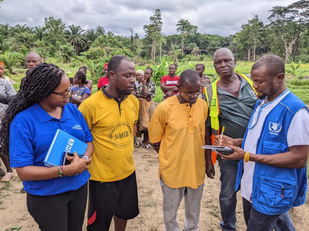 رجلان يرتديان اللون الأصفر وموظفة في هيئة الأمم المتحدة للمرأة ينظرون إلى رجل ثالث، وهو أحد موظفي برنامج الأغذية العالمي، يكتب في دفتر ملاحظات، بينما تقف مجموعة من الأشخاص في الخلفية.