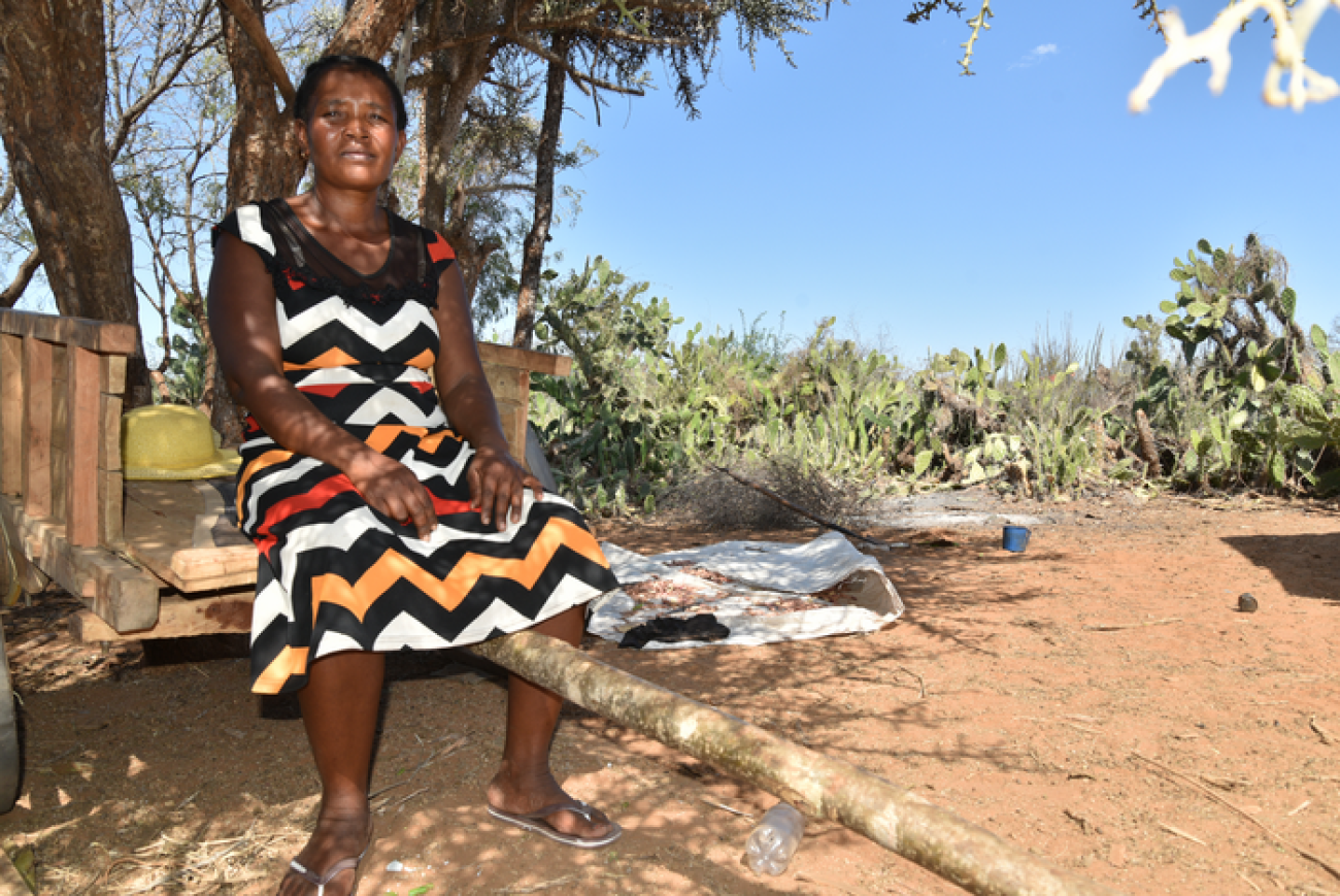 Une femme malgache vêtue d’une robe est assise sur le bord d’une brouette, à l’ombre d’un arbre, à la lisière d’un champ, et regarde la caméra, les mains posées sur les genoux.