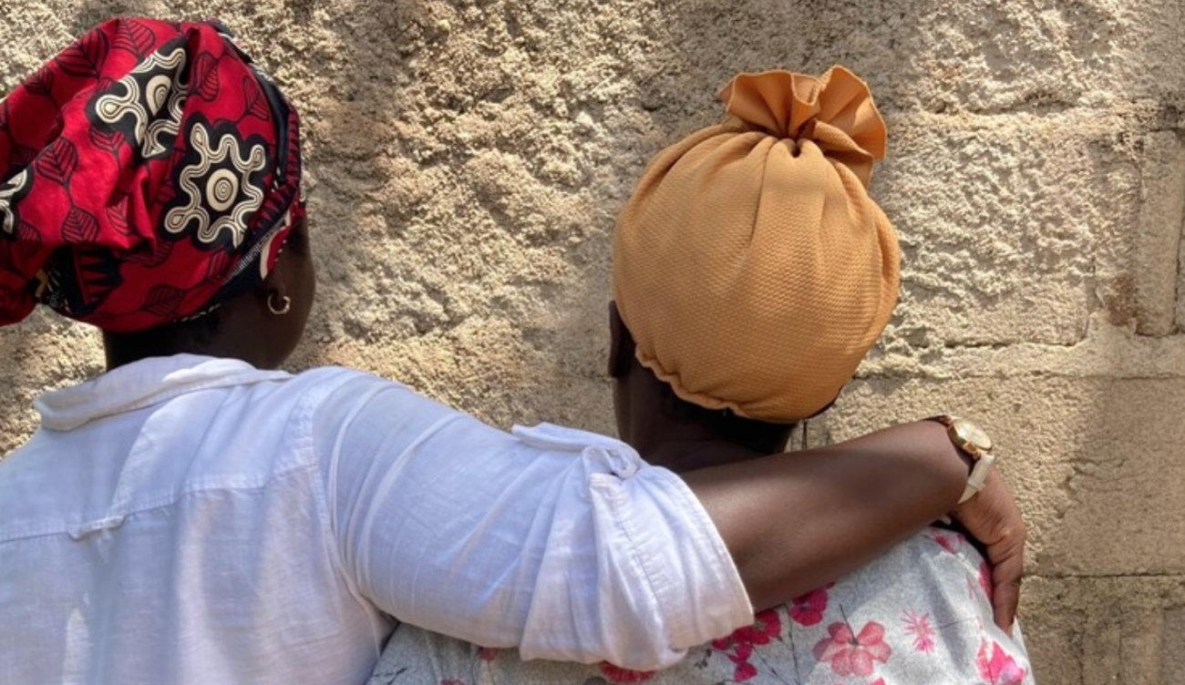 Two women in colorful headwraps embrace, and face away from the camera, in front of a tan-colored cement wall.