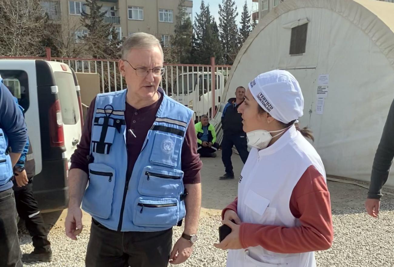 A man in a blue UN vest speaks to a rescue worker, in a white cap, amid devastation from an earthquake.