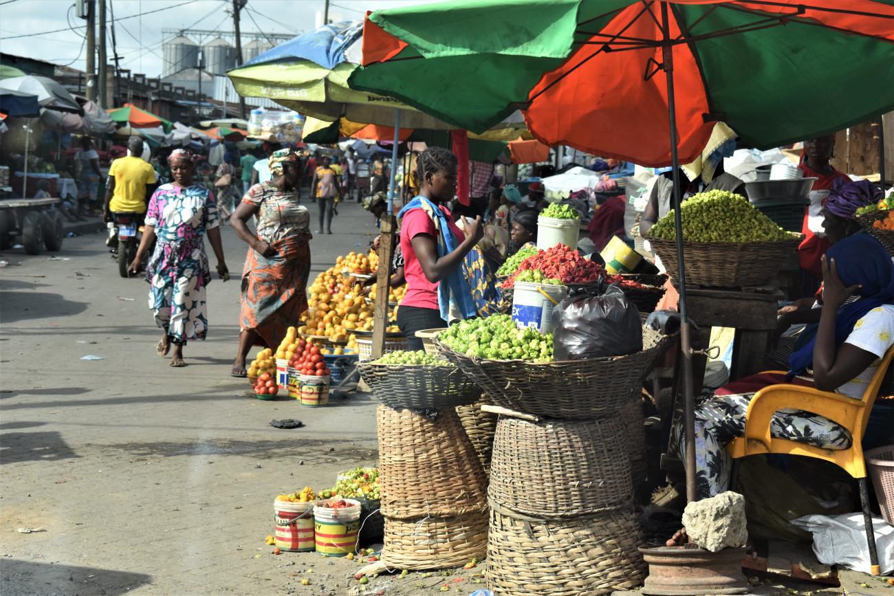 A market scene with men and women walking and surrounded by baskets with vegetables and fruits under umbrellas.