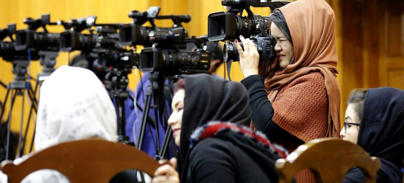Woman in a brown dress and headscarf peering beneath a journalist's camera in Afghanistan