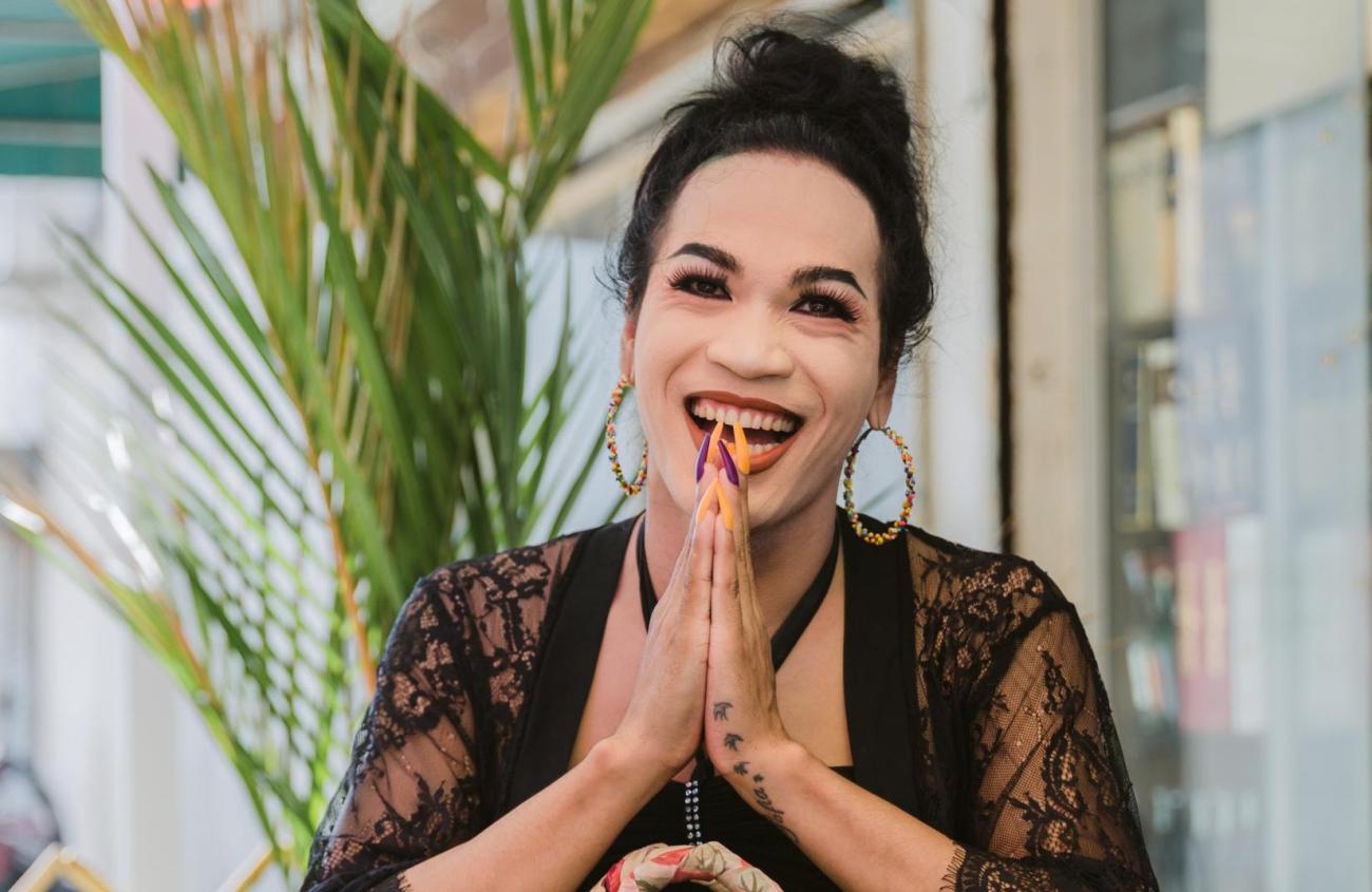A person in a black shirt folds their hand and smiles while the background has a green plant