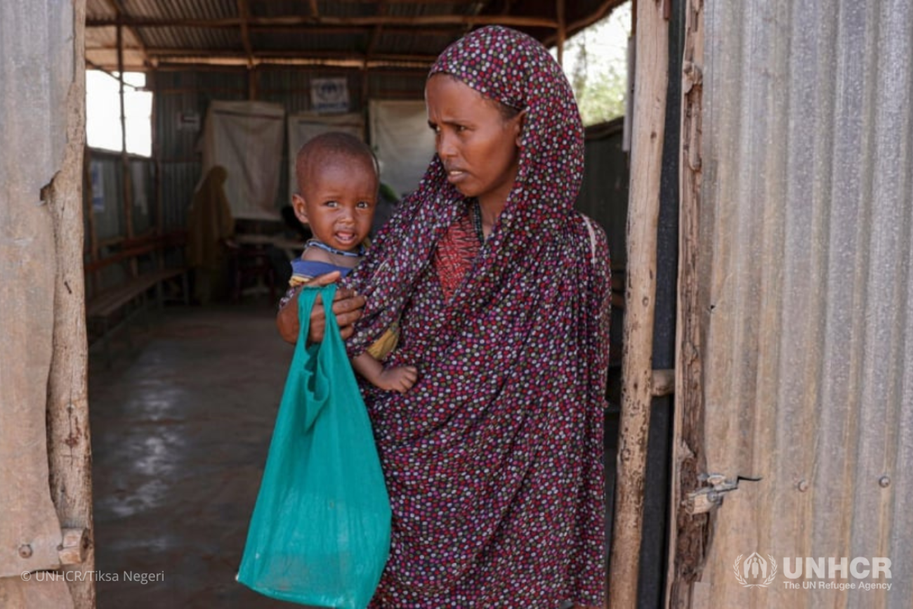 Woman in a brown dress holds a baby in blue as she stands in a doorway of a dilapidated room
