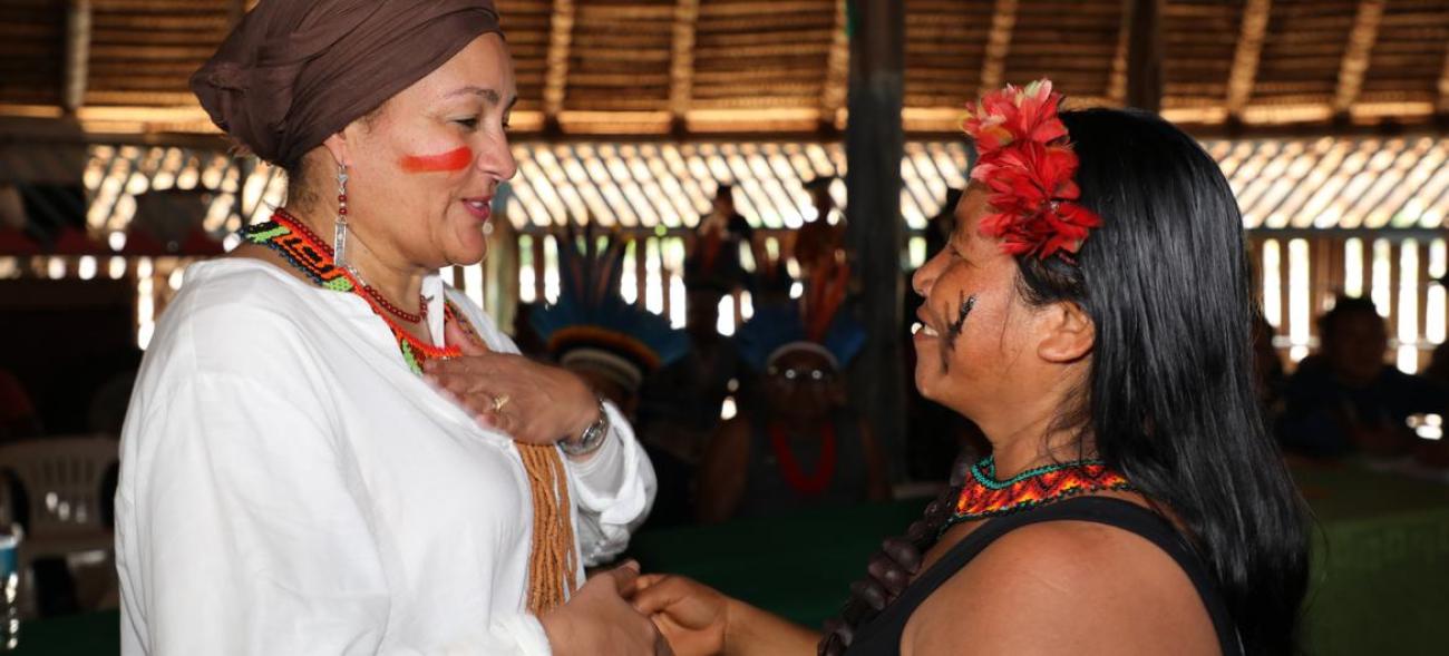 A woman in a white shirt and red headscarf shakes hands with a shorter woman in red headfeathers and face markings