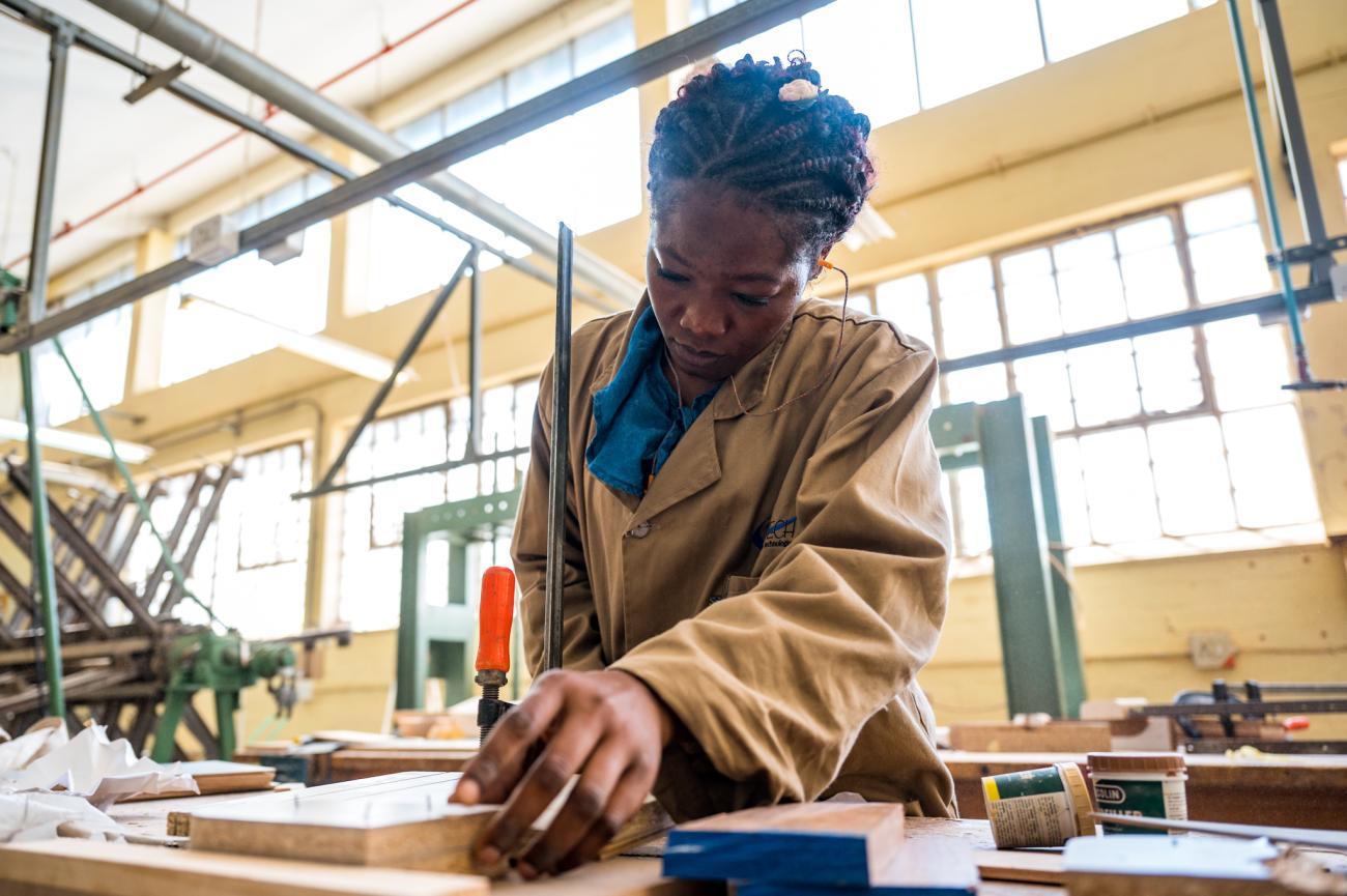 A woman in a brown shirt does woodwork on a table in a well-lit workshop