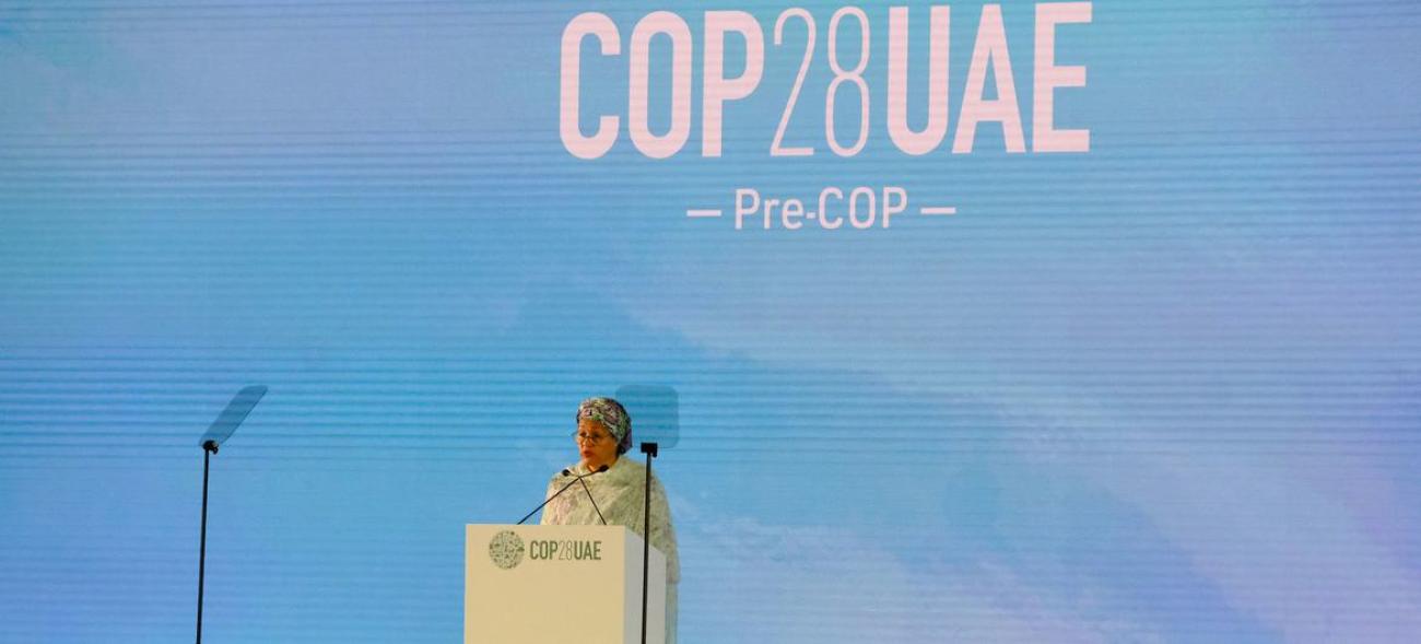 A UN official in a cream-colored outfit stands at a podium on a stage with a sign that says 'COP 28 UAE'.