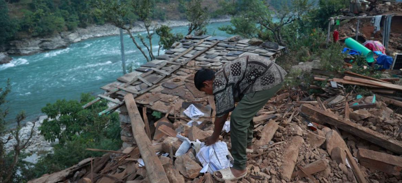 A woman in a green dress kneels over some destroyed wood right by a flowing blue river in Nepal.