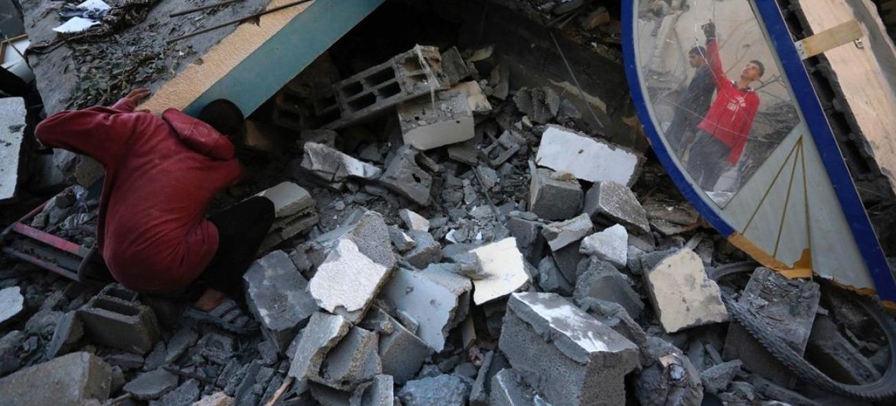 A young boy in a red shirt kneels among the rubble, stones and broken building, searching for something.