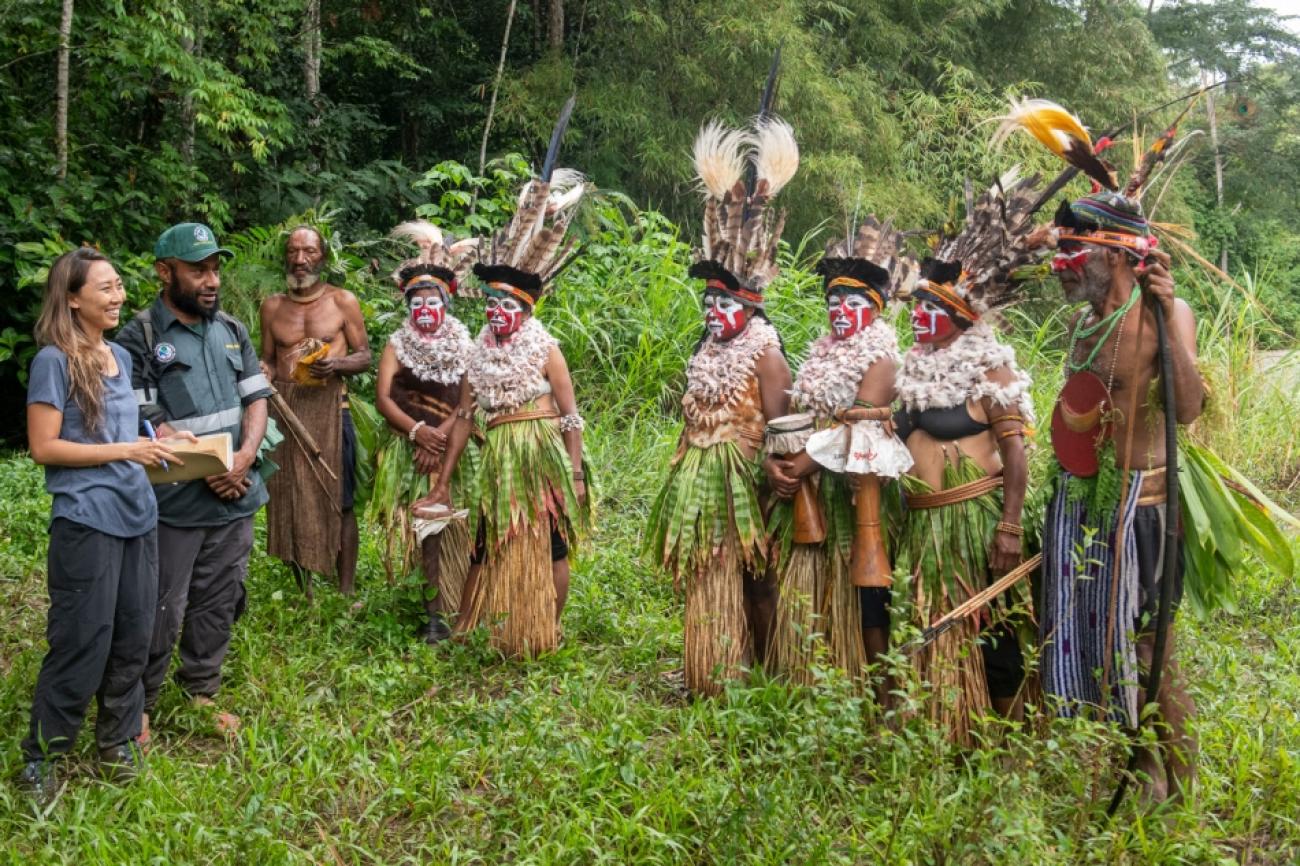 A group of indigenous peoples in traditional clothes and headgear, speak to a man and a woman dressed in dark blue attire. They are all standing in a lush green forest clearing.