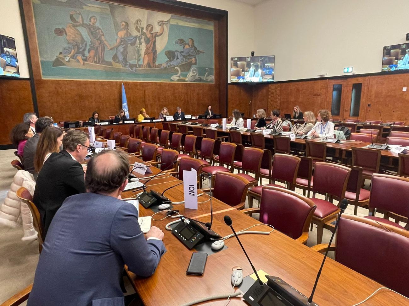 A room with people sitting up on a podium and several others in red chairs sitting around a long table