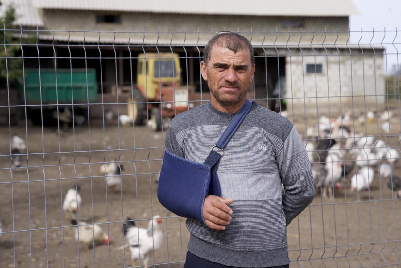 A man in a blue shirt and cast on his right arm stands in front of a grain barn and a chicken coop.