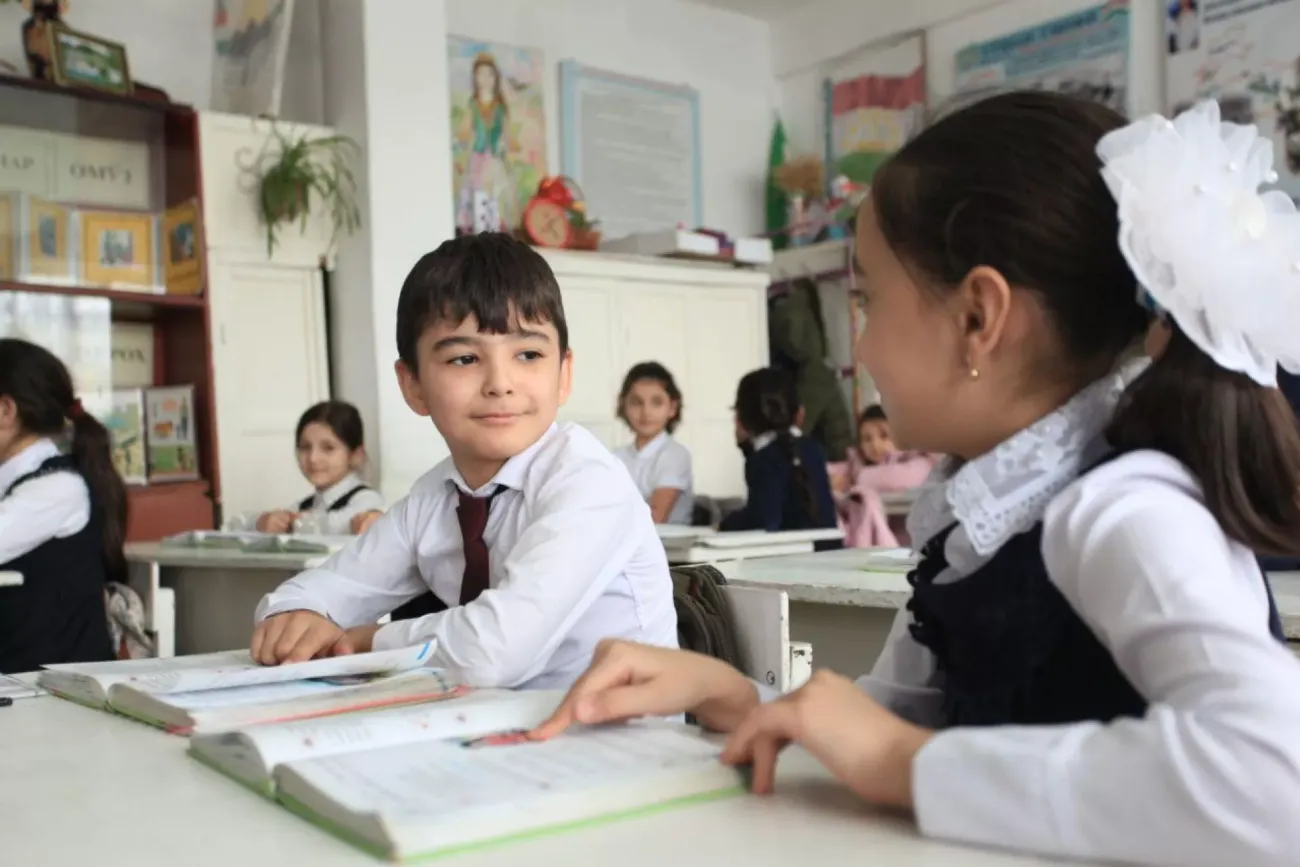 A classroom setting where a boy in a white school uniform smiles at a girl in a blue pinafore