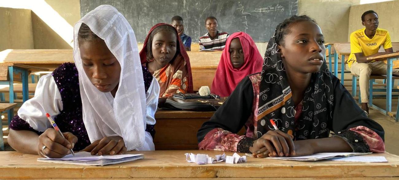 A group of four girls wearing colourful clothes sit at desks in a school, writing in their notebooks and listening to their teacher.