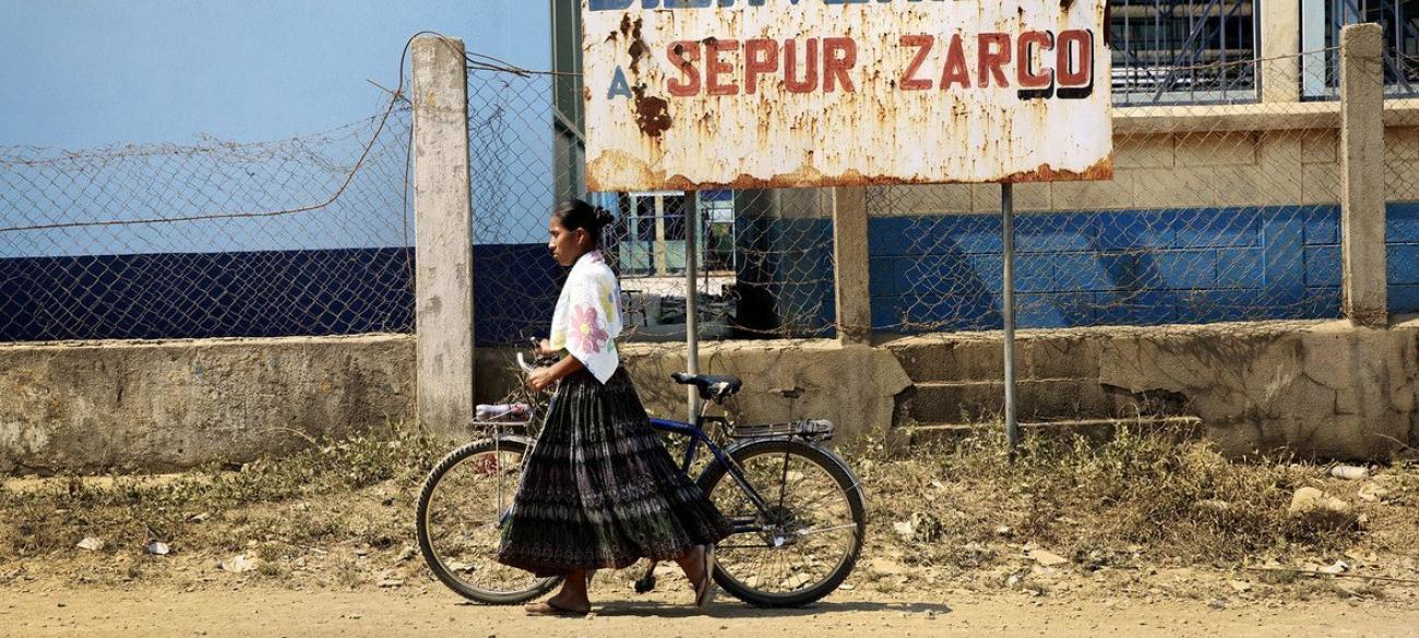 An indigenous woman walks her bicycle, there is a sign in the background and it is a dirt road. 