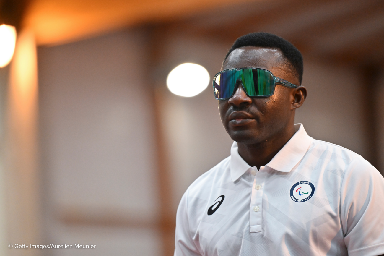 A man in a white shirt and dark visor glasses stands in a well-lit room