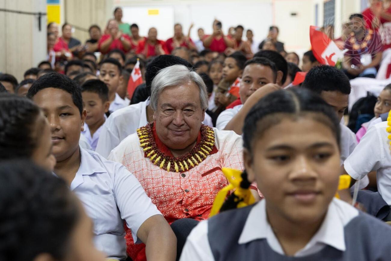 A man in a white and orange shirt and a wreath around his neck sits with a bunch of school students