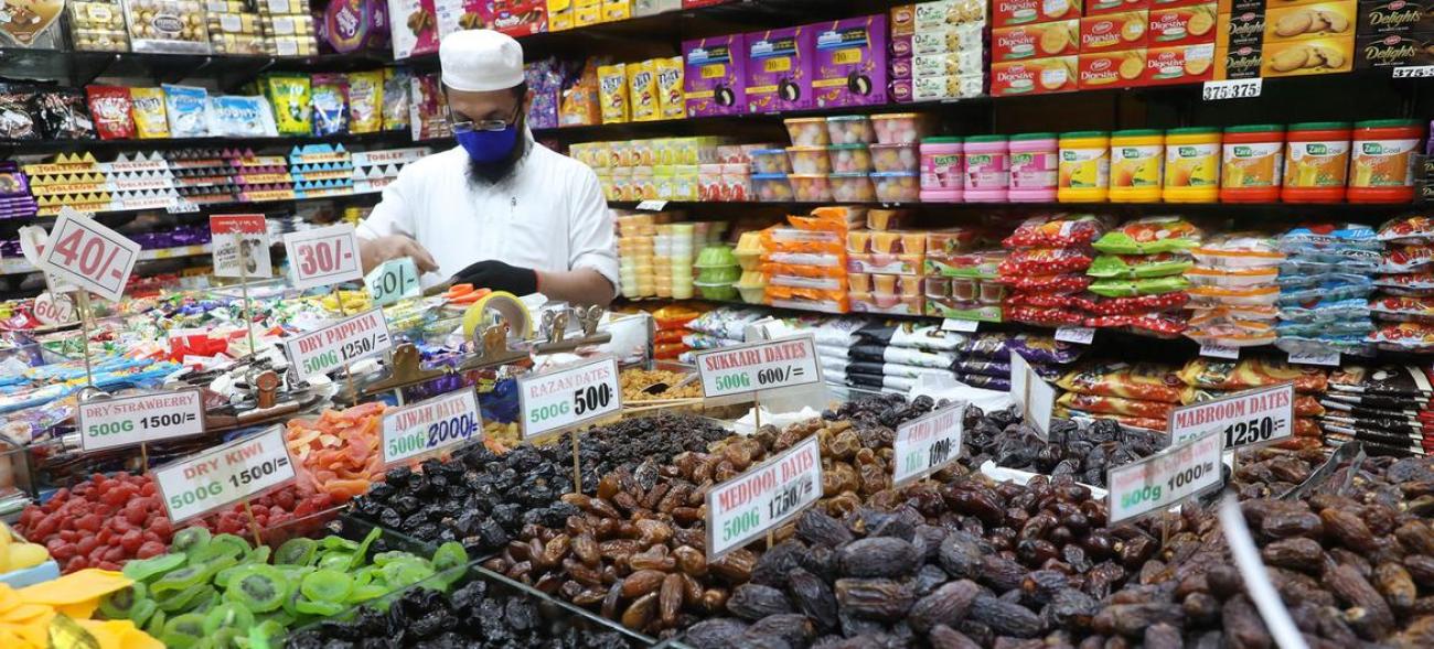 A man in a white shirt and hat, stands behind a cashier in a shop with many commodities and goods.