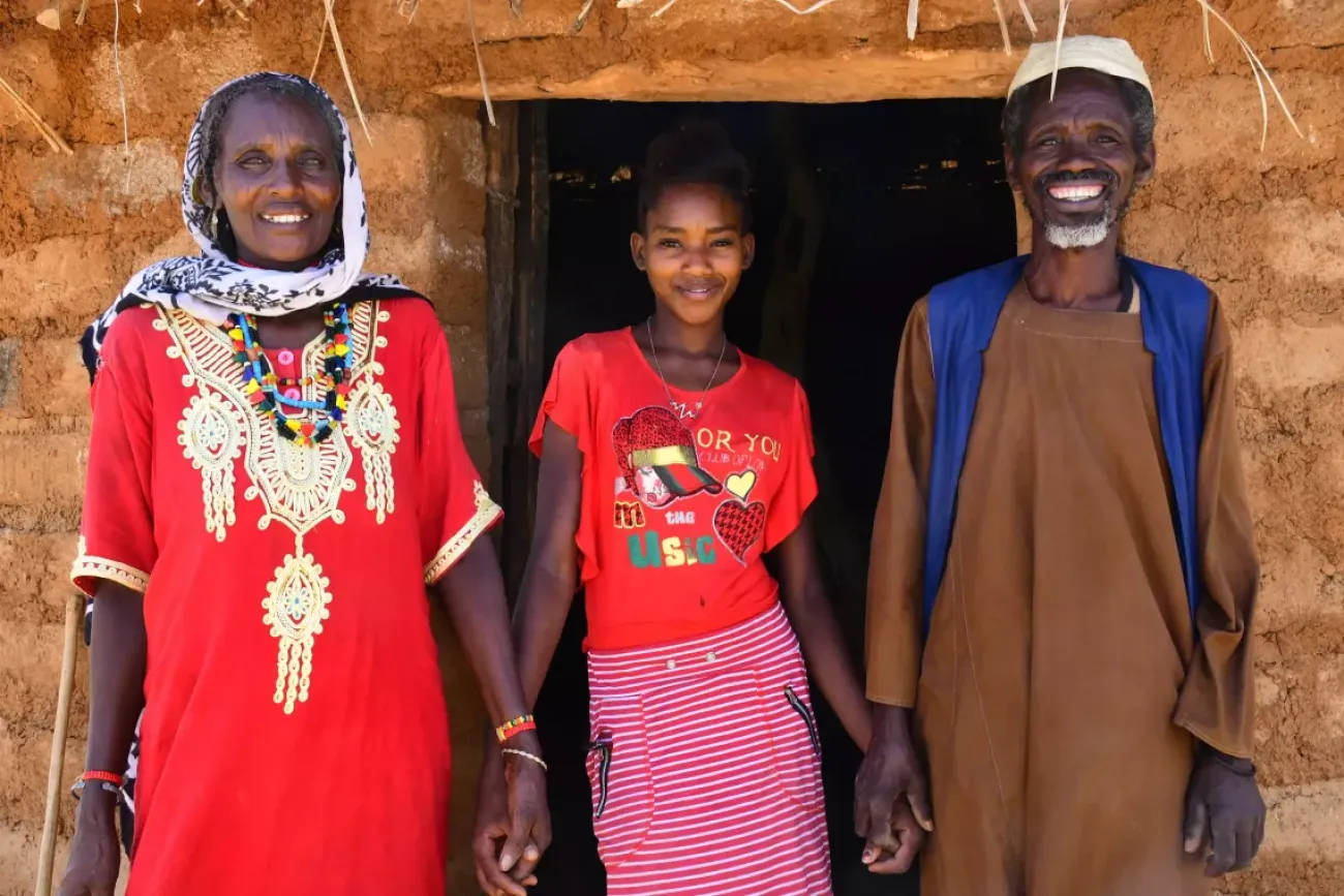 A woman, a young girl and a man stand outside a brick house. They are wearing colourful clothes and smiling.