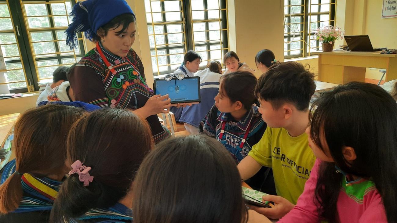 A teacher holds a tablet in front of her students in a classroom.