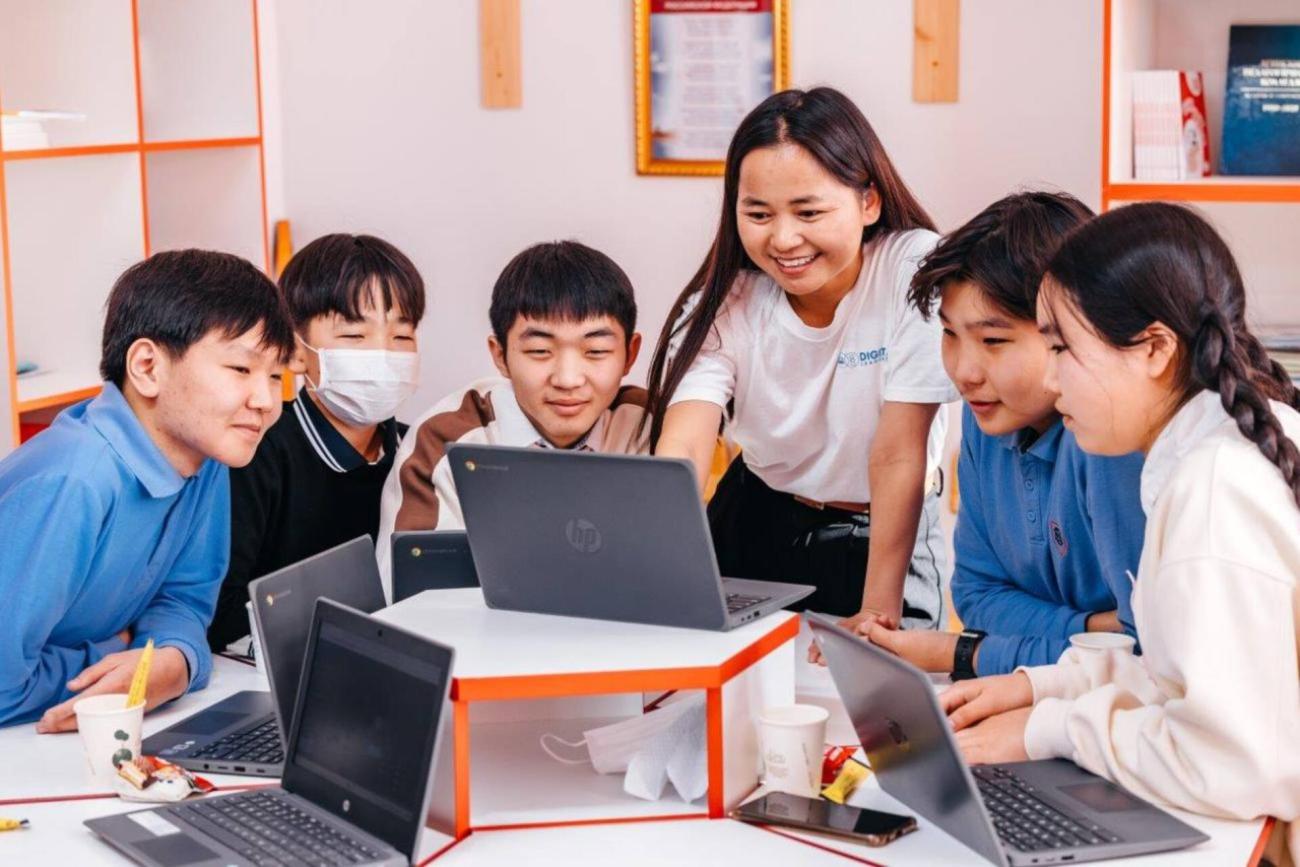 A group of smiling schoolchildren surround an instructor who is pointing at a computer screen.A group of smiling schoolchildren surround an instructor who is pointing at a computer screen.