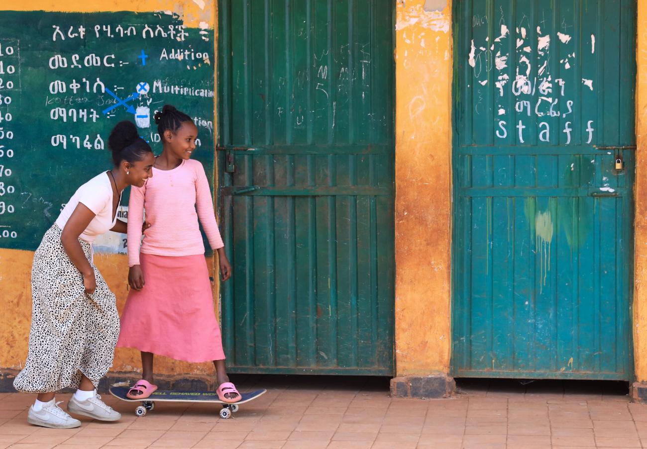 Two girls in front of a school classroom; one of the girls is on a skateboard