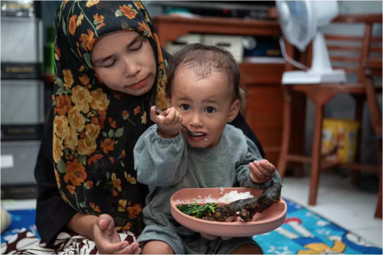A little boy is eating his food seated in his mother's lap