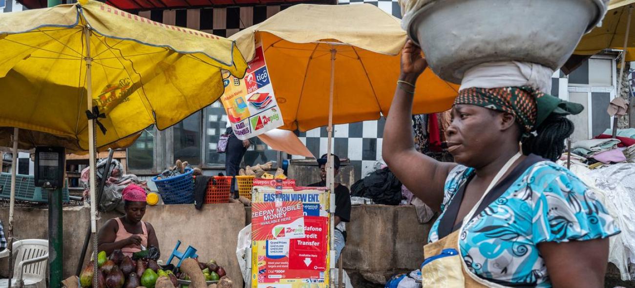 A woman holds a basket of goods on her head, in front of a produce market