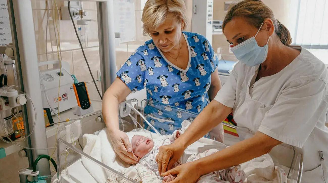 Two nurses care for a newborn child in a medical unit, connected to wires.