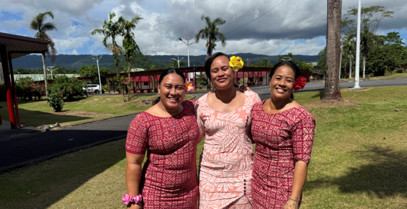Three people are standing together in colorful dresses in front of a green grassy field with trees