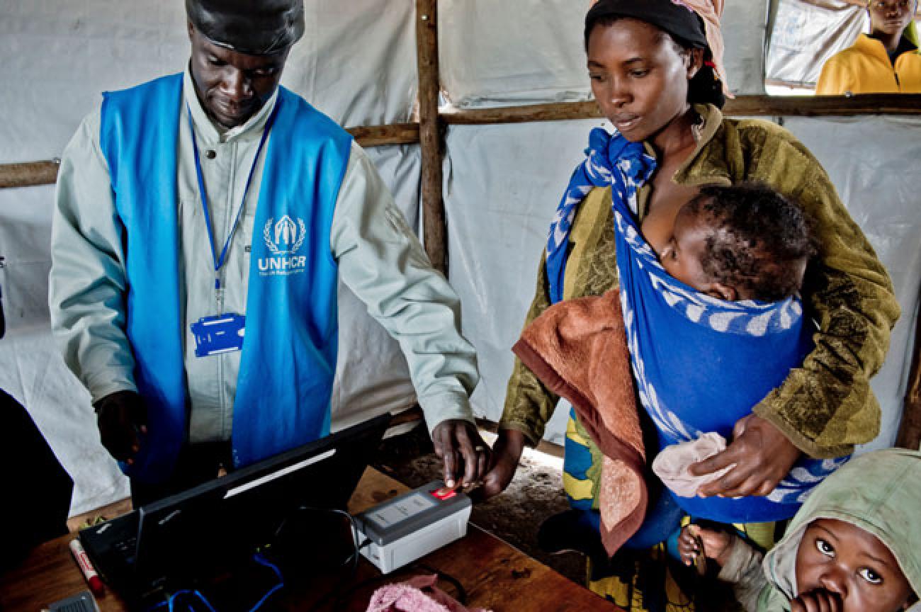 A woman holds a baby in her arms while a man in a blue United Nations vest looks at a computer.