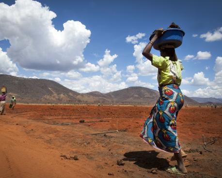 Une femme africaine traverse une plaine désertique en portant un panier sur la tête.