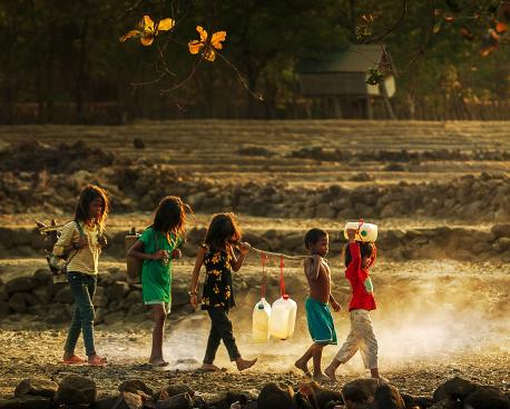 Little children walk in a line as they all hold a large stick holding jugs of water. 