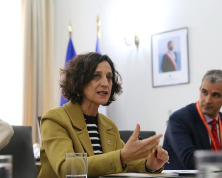 A woman in a yellow suit gestures as she talks while a man, faded in the background, listens.