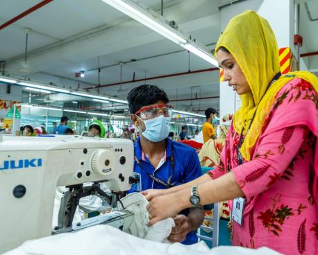 一位披着黄色头纱粉丝连体裙的女性正操作一台纺织机器，旁边站着的是一位戴着口罩 防护眼镜的蓝色衣服的车间工人