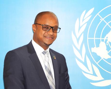A man in a charcoal grey suit with a grey tie and wearing spectacles stands in front of blue wall with a white UN logo