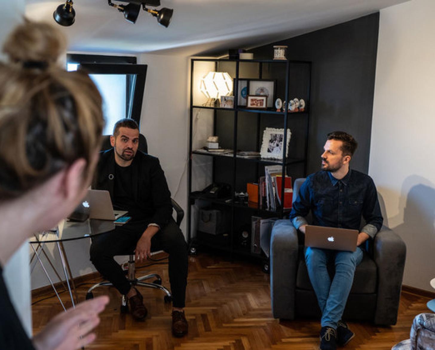 Two men hold their laptops while conversing with a woman and each other