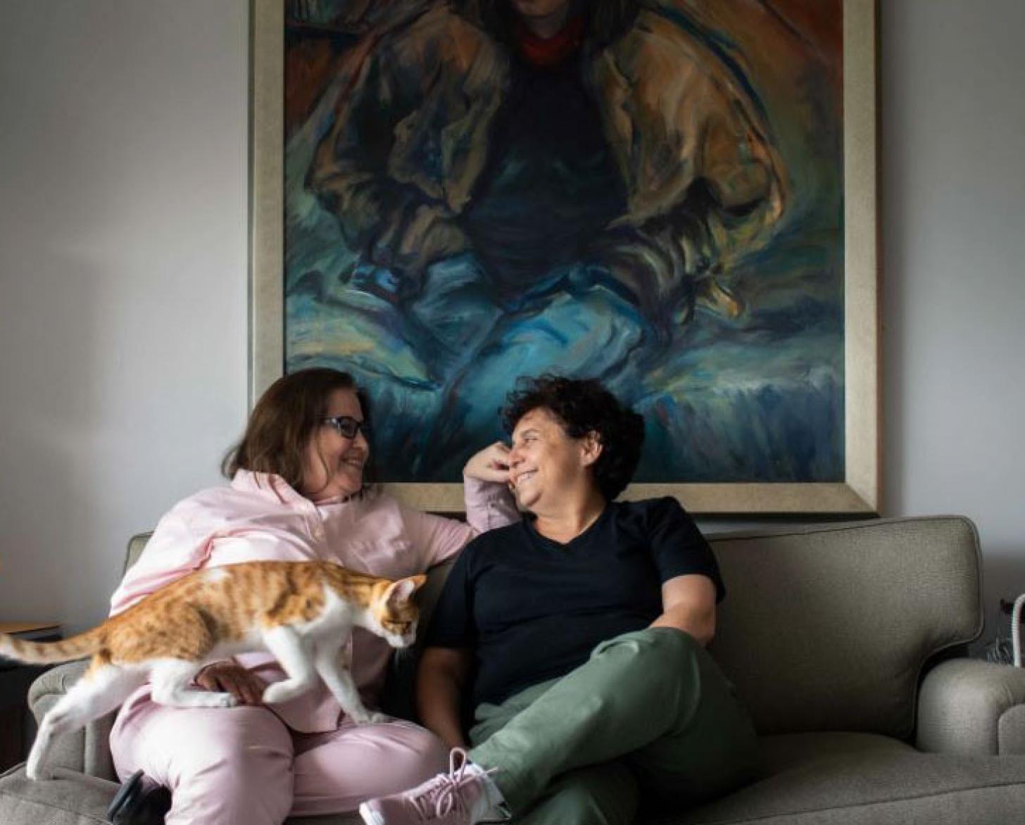 Susel and Gracia lovingly look at each other and smile, while sitting on a couch with a cat making its way to their lap.
