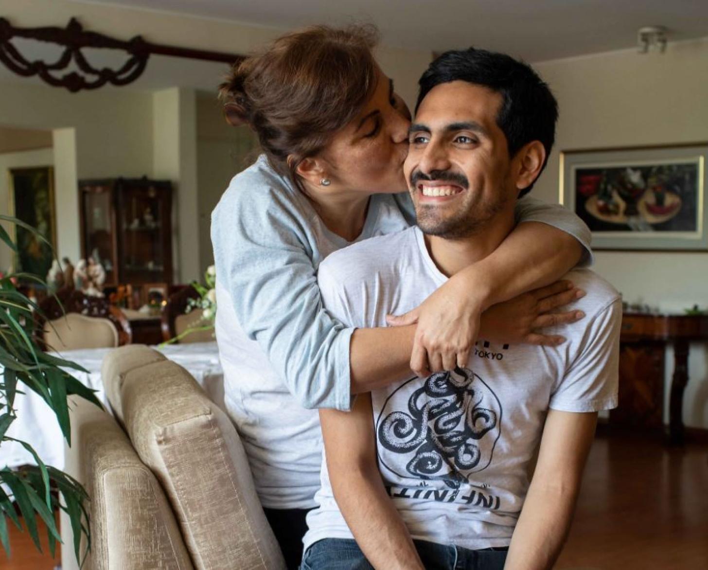Cecilia, mother, wraps her arms around her son, Carlos, while she kisses his cheek and he smiles. 