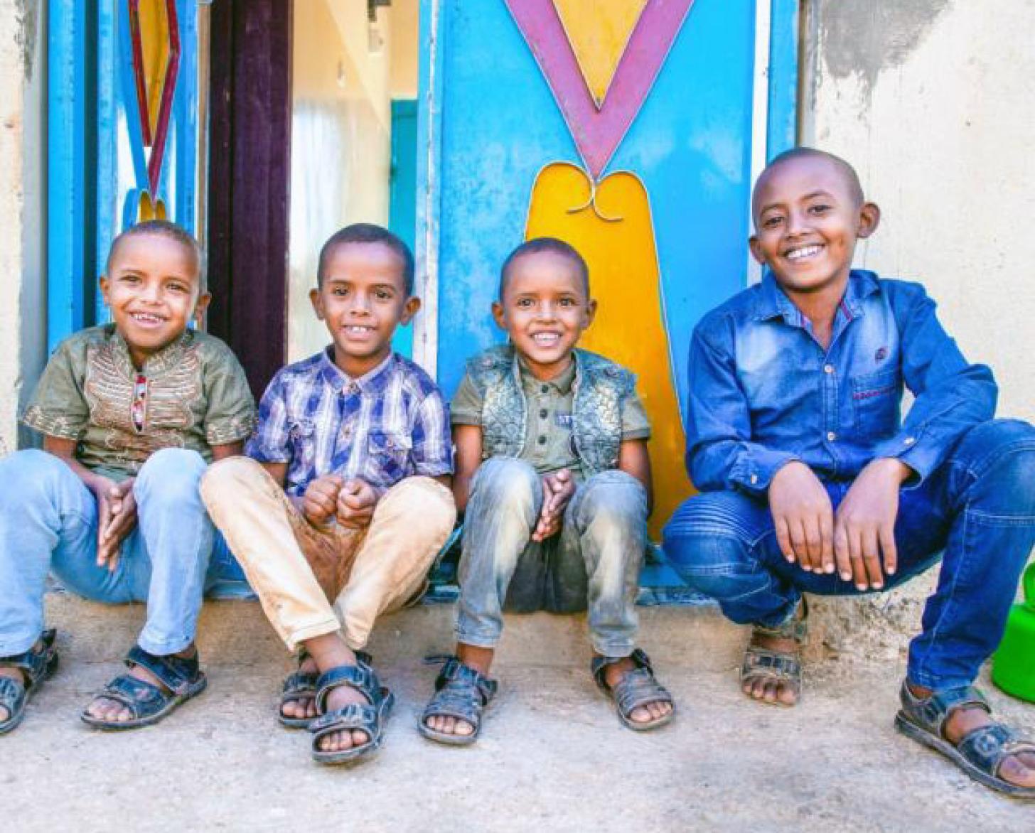 Four young boys sit outside on the step in front of an entrance. They all smile happily at the camera. 
