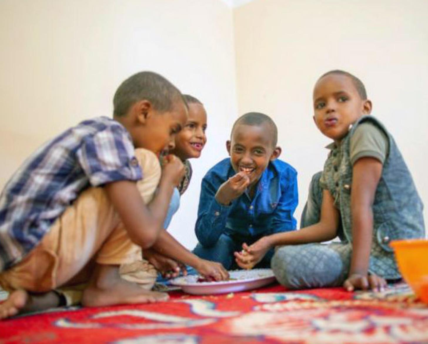 Shows a photo from a low angle of the boys sitting around a plate of food, eating and smiling at the camera and each other. 